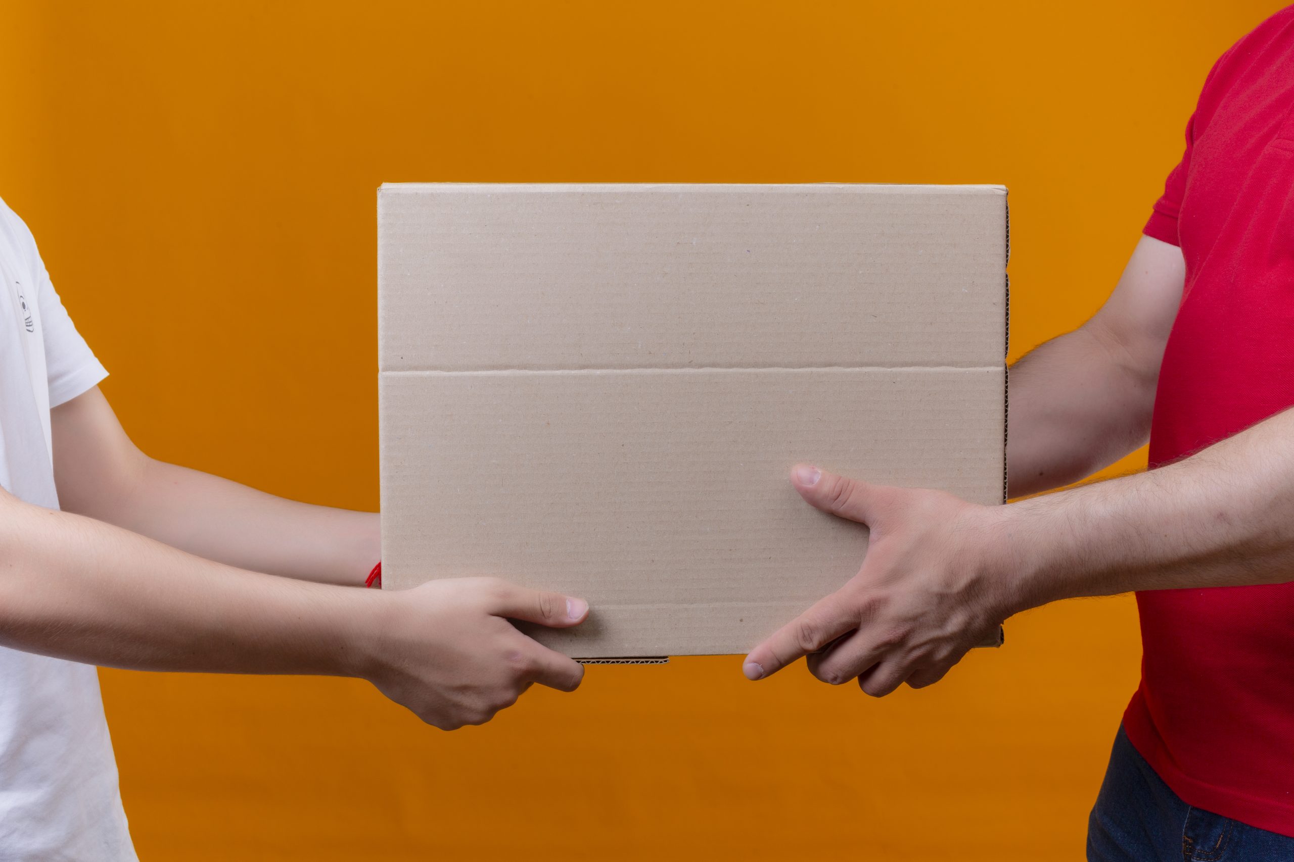 cropped view of delivery man in red uniform giving box package to a customer over isolated orange background